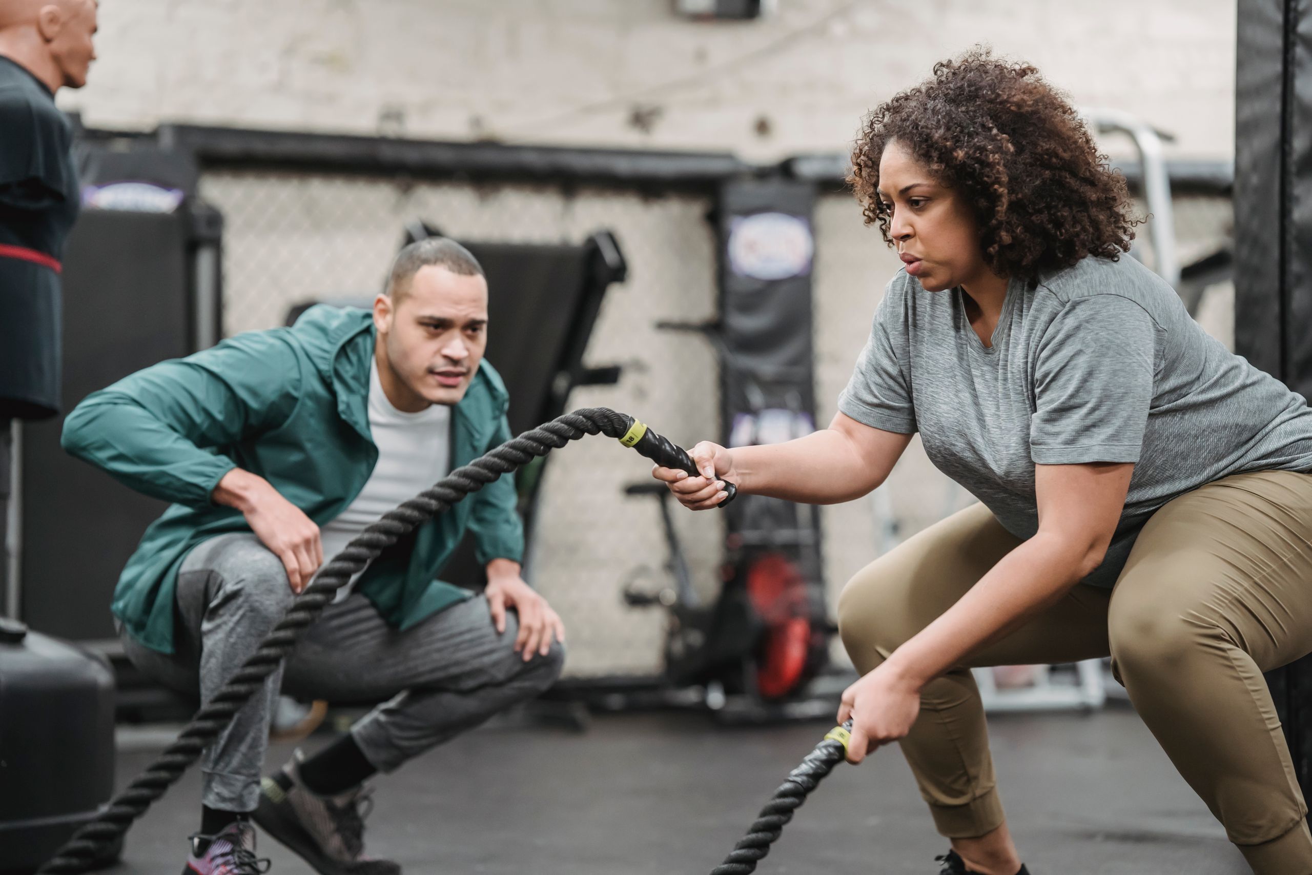 Concentrated plus sized African American female in sportswear exercising with battle ropes near fit male trainer in modern sports club